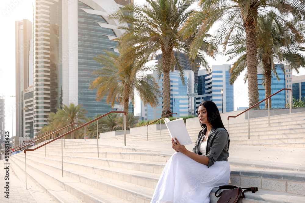 Close up portrait of young woman seating on stairs and reading a book in a nice sunny summer day in center of Doha. Concept of recreation, education and study, time out Doha, leisure time