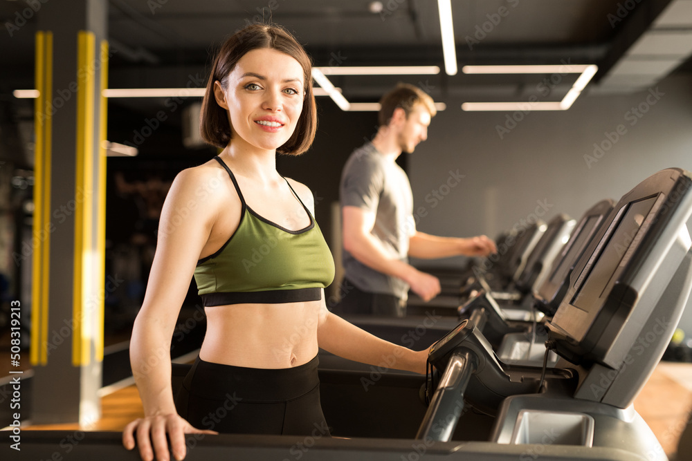Fototapeta premium Fit young woman in sportswear looking at camera while training on treadmill with young man in the background
