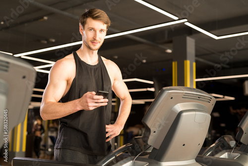 Young male athlete standing and using his smartphone before training on treadmill in gym