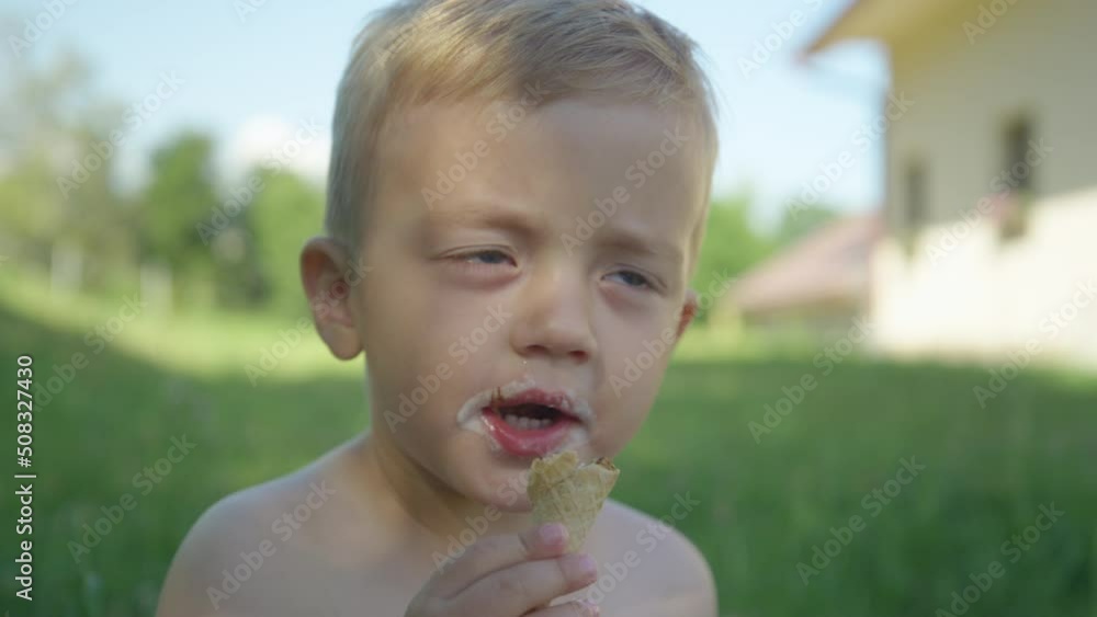 CLOSE UP: Cute little boy eating ice cream cone in garden shadow on a summer day. Adorable toddler enjoying vanilla ice cream cone in shady part of backyard. Complete refreshment on a hot summer day. CLOSE UP: Cute little boy eating ice cream cone in garden shadow on a summer day. Adorable toddler enjoying vanilla ice cream cone in shady part of backyard. Complete refreshment on a hot summer day.