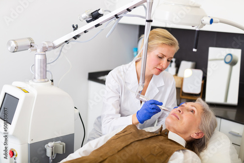 Professional female beautician making procedure for laser resurfacing of aged female client face skin on modern equipment in clinic of aesthetic cosmetology