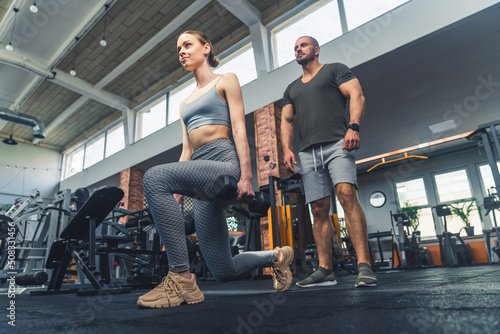 Fototapeta Naklejka Na Ścianę i Meble -  Brunette european fit woman in her 20s doing exercise at a gym, leaning one of her knees on the floor. Male muscular personal trainer watching her closely. High quality photo