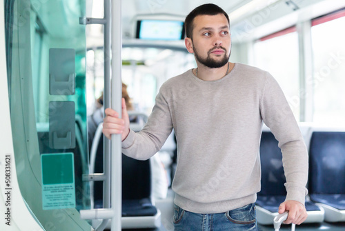 Young bearded man traveler riding in modern streetcar, standing leaning on suitcase and holding on to handrails ..