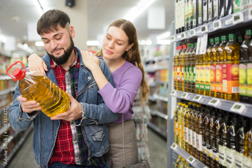 Positive young couple purchasers buying products for family dinner in supermarket, choosing organic vegetable oil