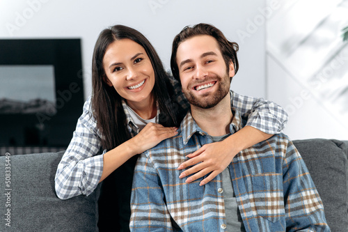 Portrait of happy caucasian couple, man and woman spending time together, hugging at home seated on couch in living room, looking at camera, smiling happily, harmonic relationships concept