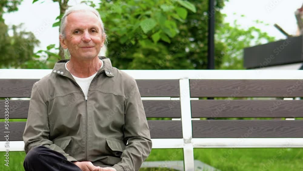 Stylish elderly man with long hair smiling on bench in public city park. Happy senior man looking at camera Stylish elderly man with long hair smiling on bench in public city park. Happy senior man looking at camera
