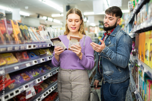 Young positive couple shopping together in supermarket, choosing tasty chocolate