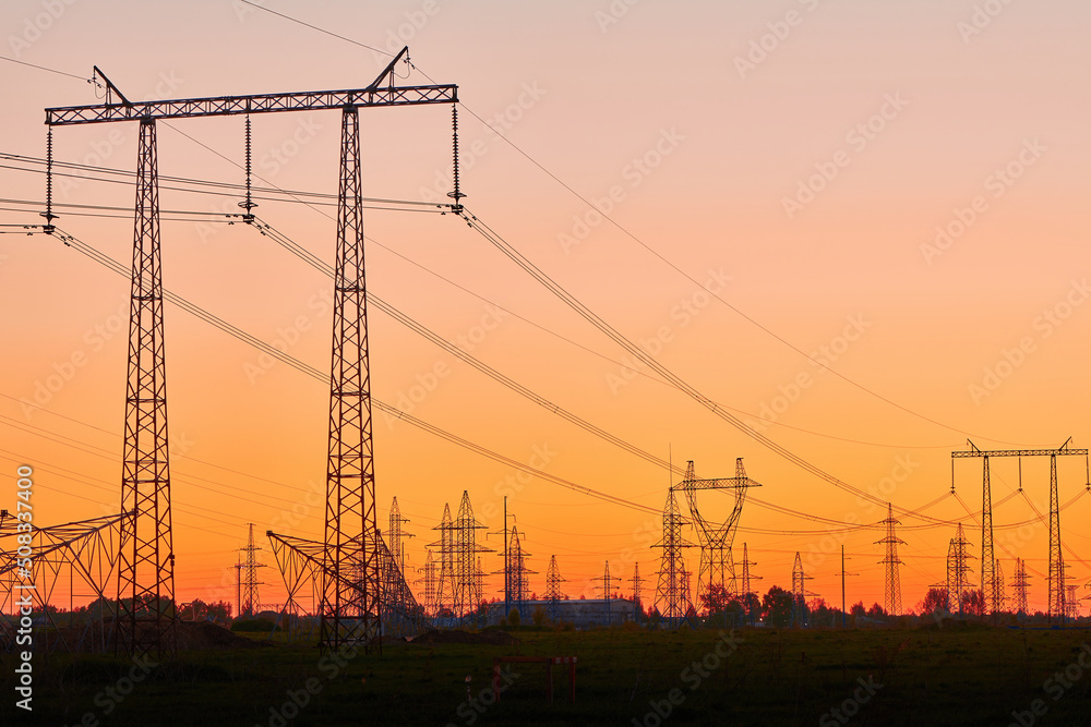 High voltage electricity towers in field at sunset and clear blue sky. Dark silhouettes of repeating power lines on orange sunrise. Electricity generation, transmission, and distribution network
