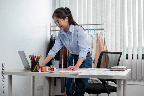 Beautiful young Asian fashion tailor with textile sewing accessories and entrepreneur designer sketches are full of bright colors on the desk with a laptop for creative ideas in the studio