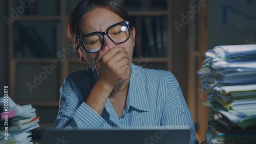 Overworked young Asian office employee wearing eyeglasses yawn during working on laptop computer overtime at night in office photo