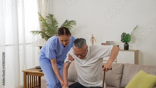 Japanese woman in-home care attendant assisting senior male to stand up from the sofa at home. the senior patient using a crutch is feeling great pain in his knee joints