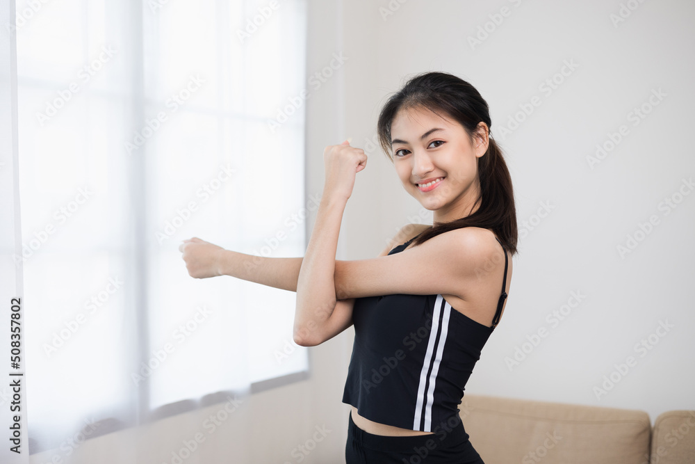 Attractive asian young fitness woman stretching arms warming up before workout at home. Beautiful smiling Female wearing sportswear exercise training yoga in living room.