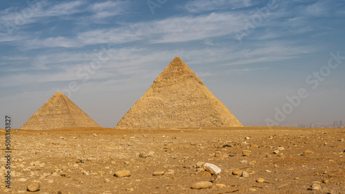 The great pyramids of Cheops and Chephren against the blue sky and clouds. The stones are scattered on the sandy soil of the Giza plateau. Silhouettes of Cairo buildings in the distance. Egypt