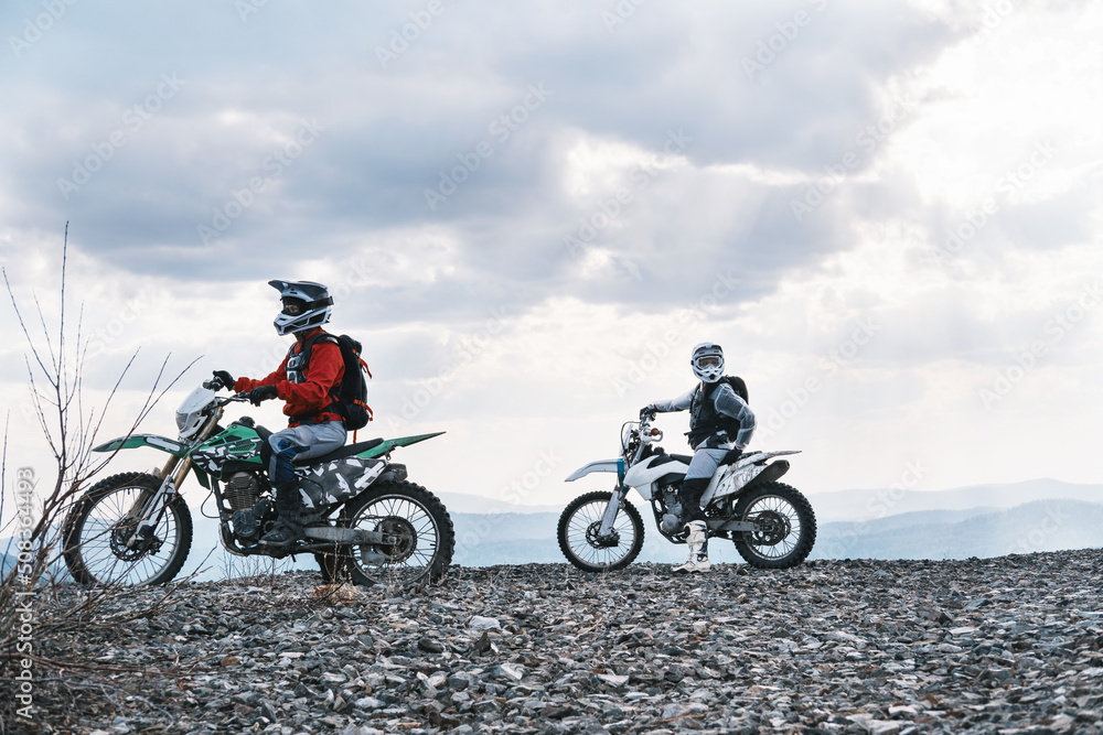 Couple of motorcyclists traveling offroad, standing on mountain top, enjoying view Couple of motorcyclists traveling offroad, standing on mountain top, enjoying view