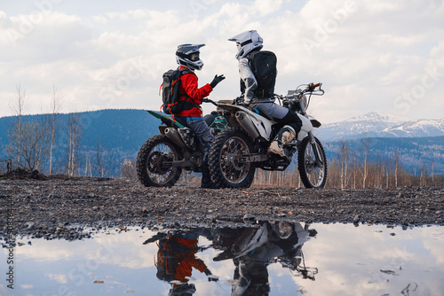 Females wearing helmets and motorcycle gear sitting on motocross motorcycles discuss the journey. Reflection in puddles, snowy peaks on horizon. Offroad travel © Annatamila