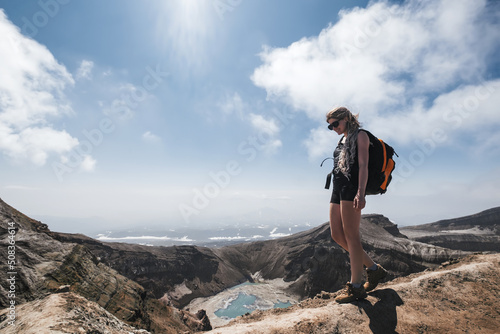 Female traveler wearing sun glasses and backpack walking on crater of volcano above blue lake. Gorely volcano, Kamchatka photo