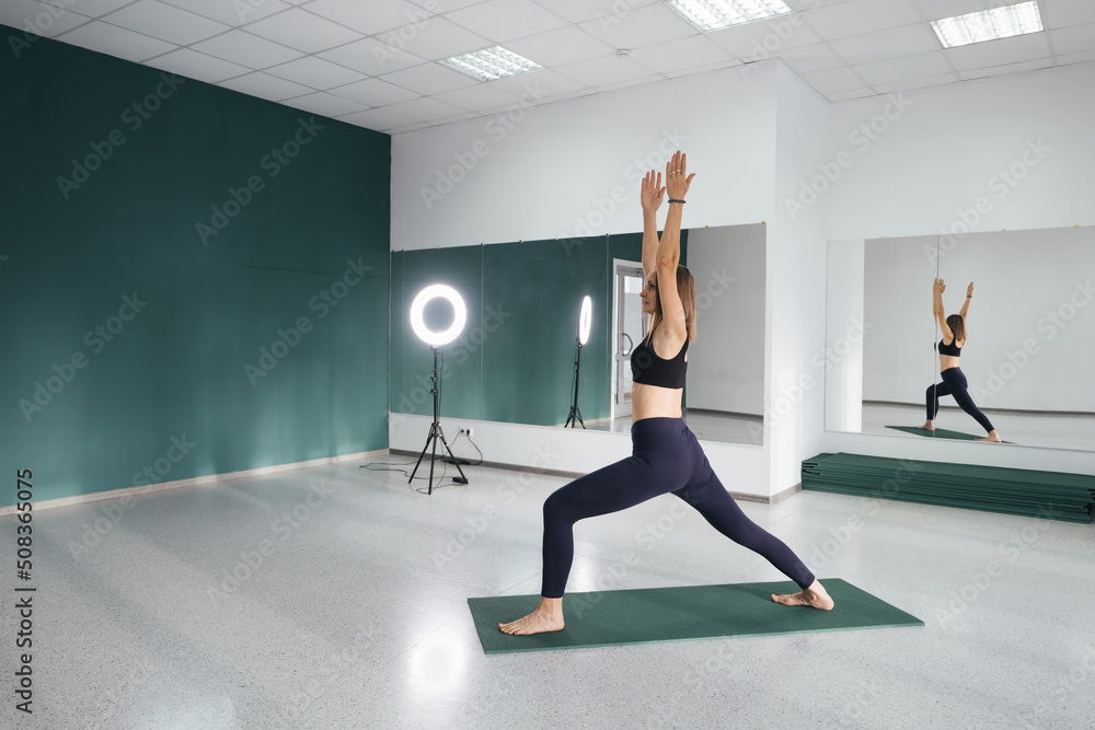 Attractive female middle age 50 years old doing standing hero yoga pose near big mirror in empty yoga studio Attractive female middle age 50 years old doing standing hero yoga pose near big mirror in empty yoga studio