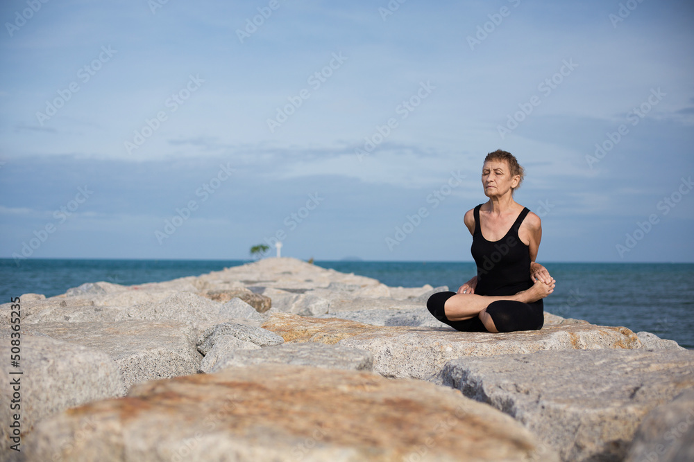 Elderly female sitting in closed lotus position meditating with closed eyes on blue sea beach