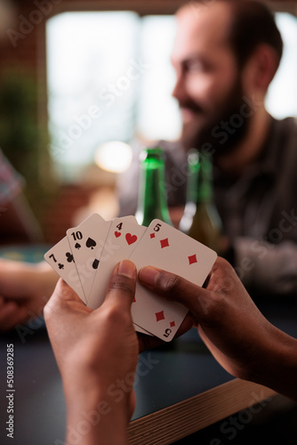 Close up shot of african american hands holding cards while playing with friends at home. Diverse group of people sitting at home in living room while relaxing with snacks, beer and society games.