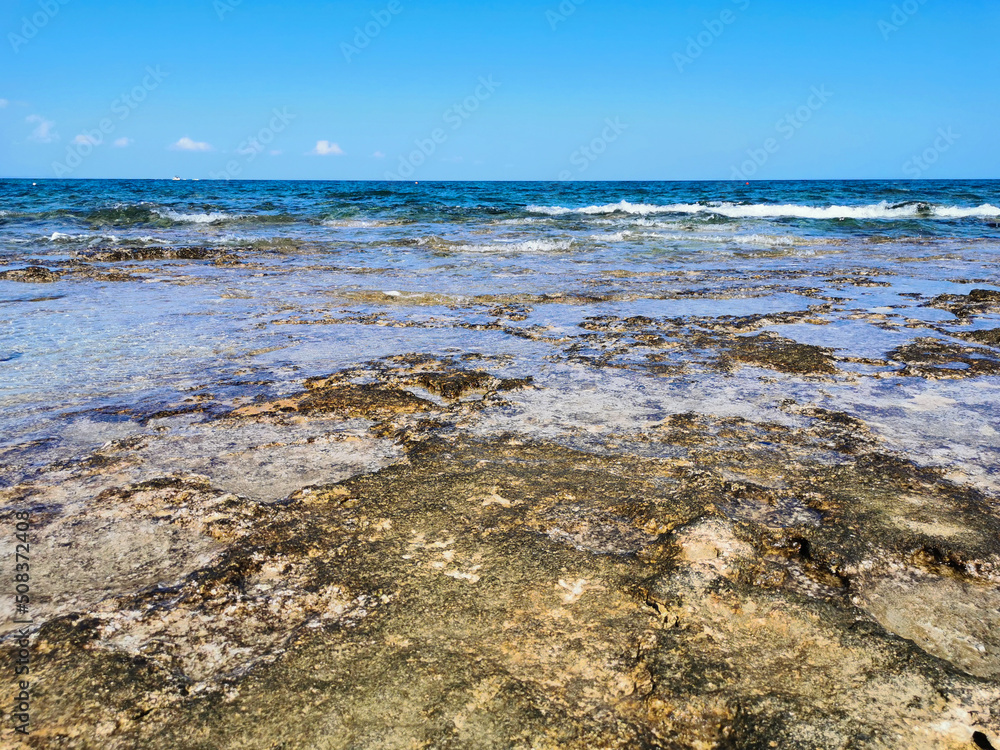 The coast of the Mediterranean Sea, long frozen lava, in the recesses of which there is sea water against a blue sky with clouds.