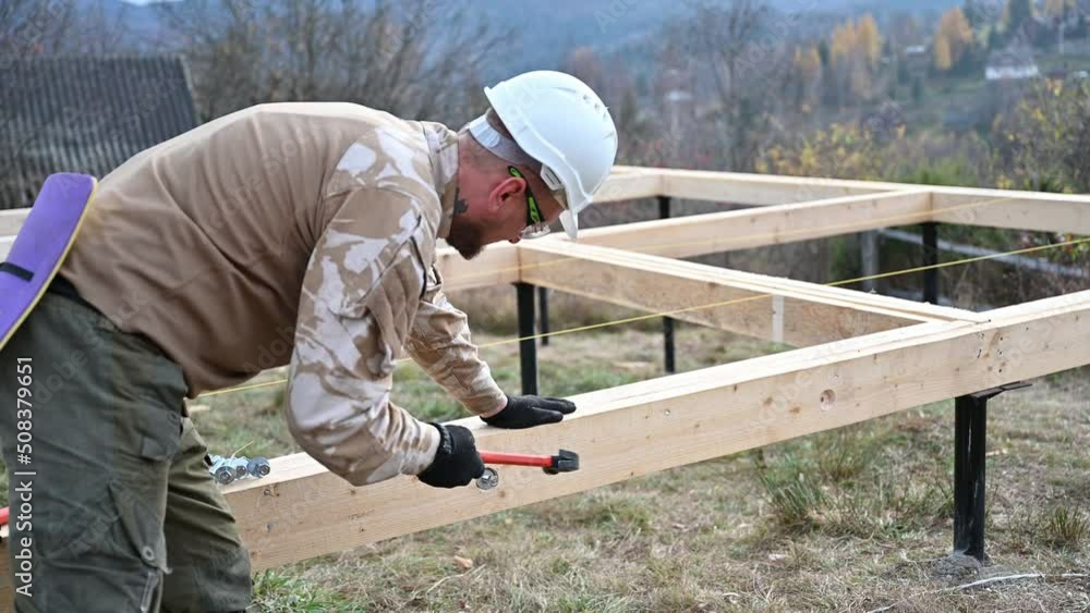 Man worker building wooden frame house on pile foundation. Carpenter hammering bolt into wooden truss, using hammer. Carpentry concept. Man worker building wooden frame house on pile foundation. Carpenter hammering bolt into wooden truss, using hammer. Carpentry concept.