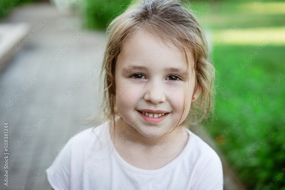 Child's portrait girl in light T-shirt close-up against background green grass smiling sweetly and looking at the camera. Caucasian little girl Child's portrait girl in light T-shirt close-up against background green grass smiling sweetly and looking at the camera. Caucasian little girl