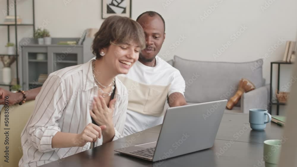 Medium shot of young happy multiethnic couple watching funny dancing videos on laptop and repeating movements, having fun while spending leisure time together at home Medium shot of young happy multiethnic couple watching funny dancing videos on laptop and repeating movements, having fun while spending leisure time together at home