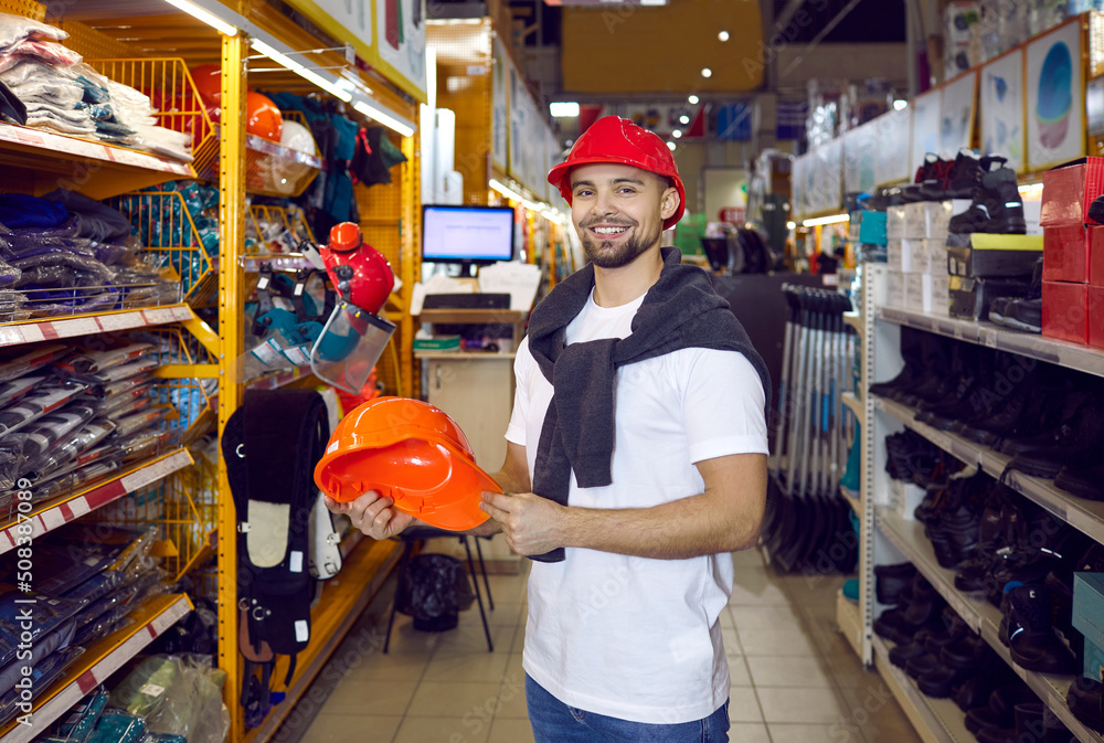 Shopping in hardware store. Man during shopping chooses protective hard hat in hypermarket of building materials. Portrait of smiling satisfied male shopper holding hard hat standing in aisle of store