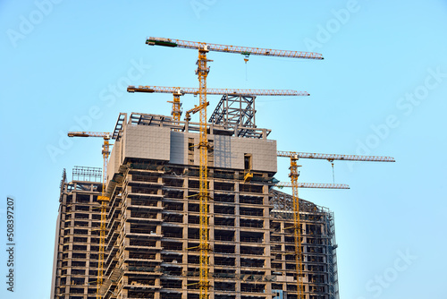 Top of three modern skyscrapers under construction with four yellow tower cranes along them in the process of installing windows on concrete structures against a clear blue sky in sunny weather
