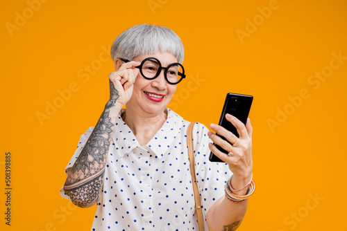 Portrait of attractive funky asian tattoo cheerful grey-haired elder old senior woman touching spectacles and smartphone device internet conected isolated over yellow pastel background color