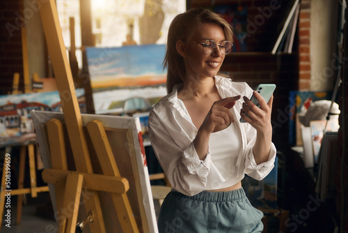 Beautiful young blonde, stands, painting with a palette at the artist's easel, joyful, uses the phone, selfie. call. art studio