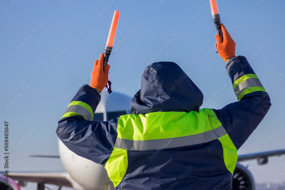 An airliner is taxiing into a parking lot at an airport, an air marshall with batons indicates a parking spot, an aeronautical engineer helps pilots park the plane An airliner is taxiing into a parking lot at an airport, an air marshall with batons indicates a parking spot, an aeronautical engineer helps pilots park the plane