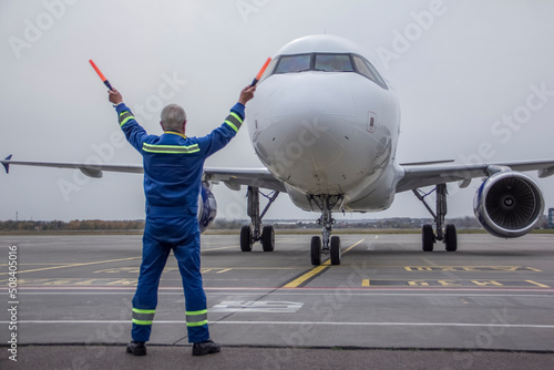 An airliner is taxiing into a parking lot at an airport, an air marshall with batons indicates a parking spot, an aeronautical engineer helps pilots park the plane