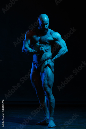 Athletic man demonstrates muscles in the light of a blue light filter on a dark background. © ksi