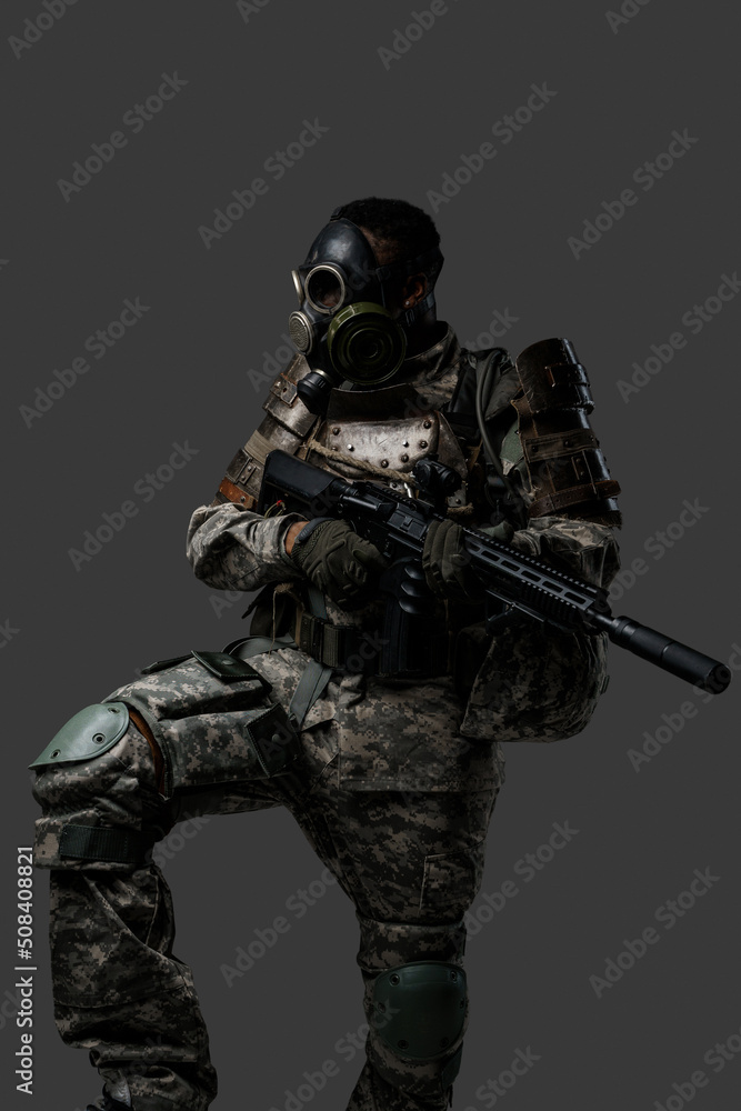 Shot of brave soldier of african ethnic with gas mask holding rifle isolated on dark background.