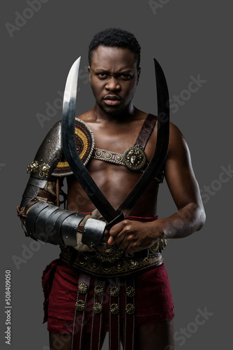 Portrait of serious african arena fighter dressed in armor holding twin swords against grey background.