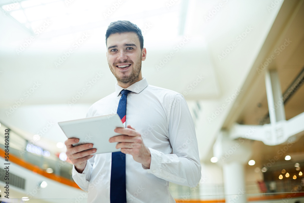 Smiling optimistic handsome young male manager with stubble standing in lobby and using digital tablet while analyzing online data