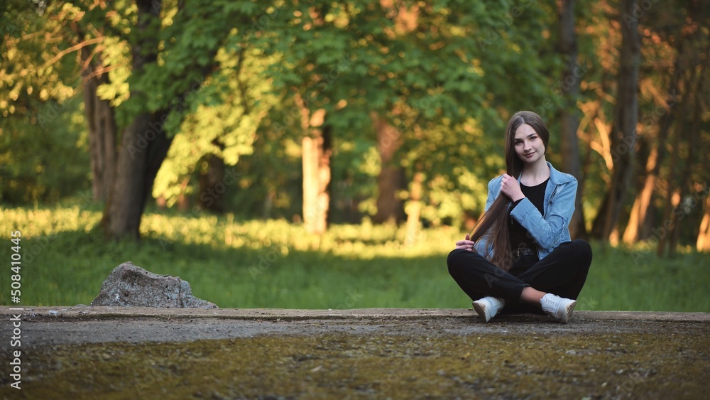 A young girl strokes her long hair while sitting in the park.