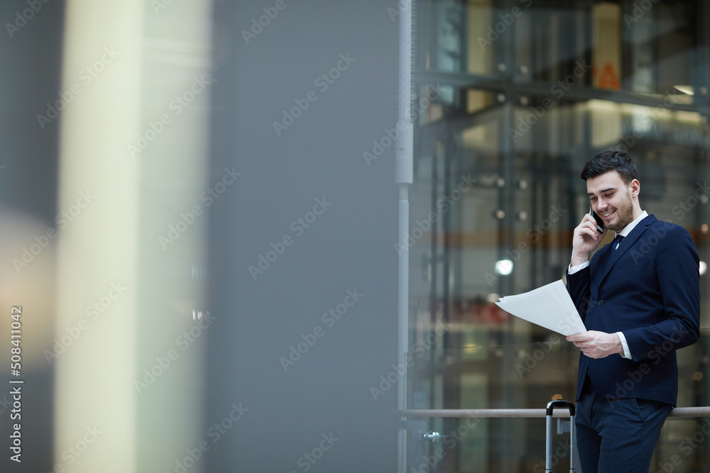 Positive handsome young male manager in suit talking by mobile phone and examining document while waiting for elevator, he has arrived in office of business partner Positive handsome young male manager in suit talking by mobile phone and examining document while waiting for elevator, he has arrived in office of business partner
