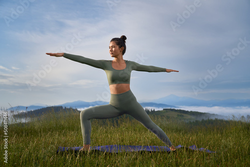 Sporty woman in activewear doing yoga exercises among summer mountains. Healthy fit lady with dark hair spending free time for outdoors workout.