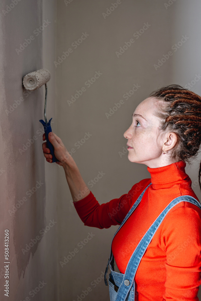 Woman with dreadlocks, in an orange turtleneck and denim jumpsuit is painting the wall with a roller in gray color. Concept of repair, renovation of the new apartment Woman with dreadlocks, in an orange turtleneck and denim jumpsuit is painting the wall with a roller in gray color. Concept of repair, renovation of the new apartment