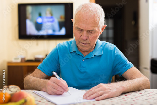 Focused senior man sitting at home reading and signing papers
