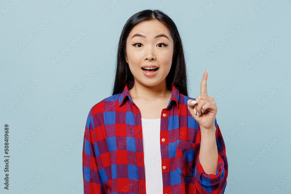Young insighted smart proactive woman of Asian ethnicity 20s in checkered shirt hold index finger up with great new idea isolated on plain pastel light blue color background. People lifestyle concept. Young insighted smart proactive woman of Asian ethnicity 20s in checkered shirt hold index finger up with great new idea isolated on plain pastel light blue color background. People lifestyle concept.