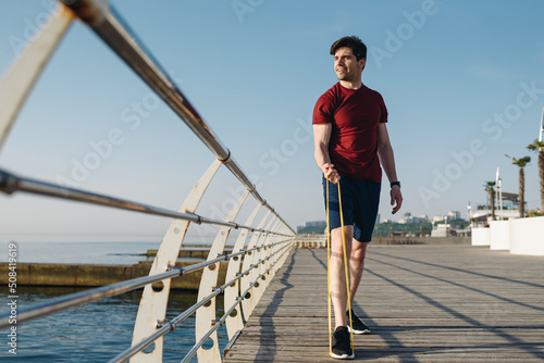 Full body young strong sporty athletic toned fit sportsman man wear sports clothes resistance bands look aside warm up training at sunrise sun dawn over sea beach outdoor on pier seaside in morning.