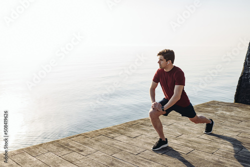 Full body side view young strong sporty athletic toned fit sportsman man in sports clothes do squates warm up training at sunrise sun dawn over sea beach outdoor on pier seaside in summer day morning.