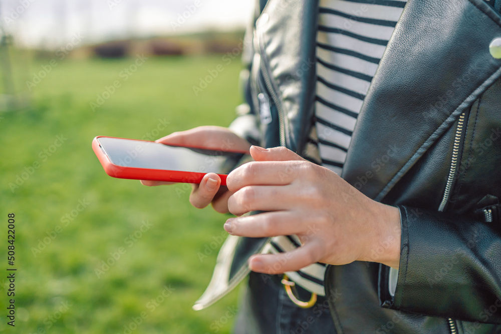 Young woman in T-shirts and stylish leather jackets texting on smartphone in the city. Close up of adult female hand using mobile phone outdoor in city park