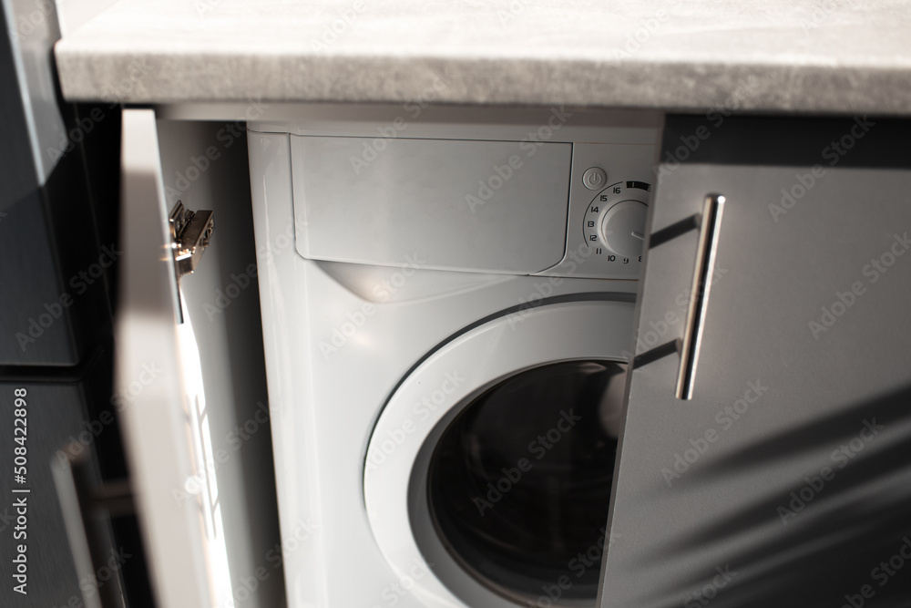 Close-up of washing machine inside kitchen cabinet with opened doors. Top view. Close-up of washing machine inside kitchen cabinet with opened doors. Top view.