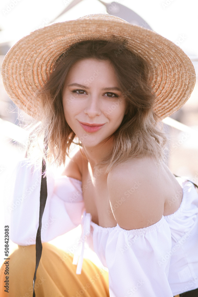 Cheerful slender lady woman girl in hat smiles broadly at the camera. Light-skinned brown-haired woman in straw hat and summer outfit holds hat