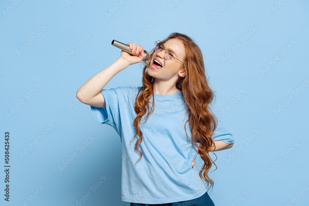 Studio shot of excited young redhead girl, student singing isolated on blue studio background. Human emotions, facial expression concept. Trendy colors Studio shot of excited young redhead girl, student singing isolated on blue studio background. Human emotions, facial expression concept. Trendy colors