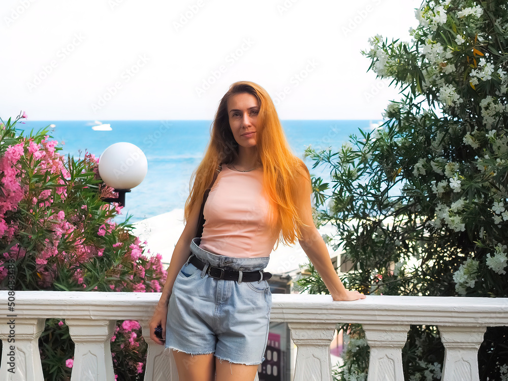 A red-haired woman stands on a white balcony against the backdrop of the sea and flowering bushes. Southern Summer Vacation A red-haired woman stands on a white balcony against the backdrop of the sea and flowering bushes. Southern Summer Vacation
